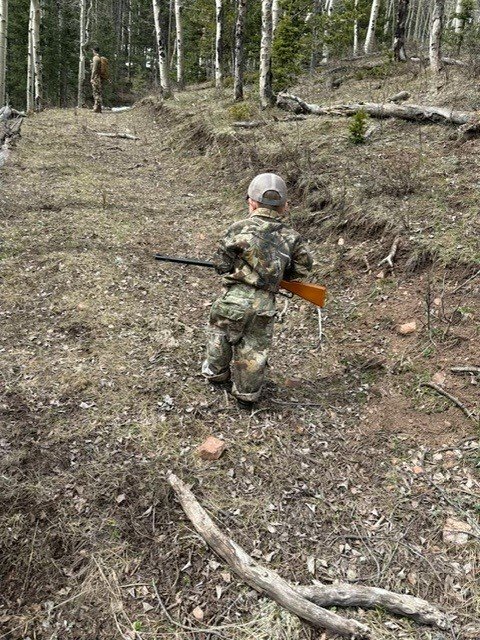 A young hunter in camo carrying a rifle through the timber at Elk Glade
